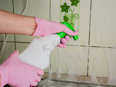 一名戴着粉红色手套的女士在燃气灶上方的瓷砖上喷洒清洁剂，以溶解油脂并清洗脏污的表面。