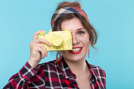 胶片格子摄影照片_一个正面美丽的年轻女子的特写肖像，她穿着格子衬衫，在蓝色背景下用黄色老式相机拍照。