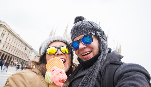 冬季旅行摄影照片_冬季旅行和意大利概念 — 快乐的年轻夫妇在米兰大教堂前用冰淇淋自拍。
