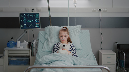 孩子住院摄影照片_住院病女儿童患者在就诊时抱着泰迪熊躺在床上休息的画像