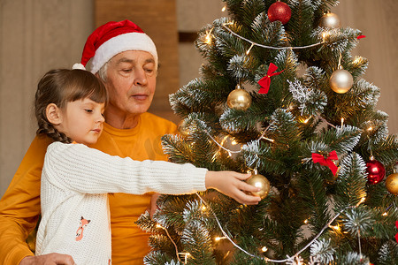 小孩圣诞老人摄影照片_老人带着小孙女在家装饰圣诞树，老人戴着圣诞老人帽子和橙色衬衫，小孩穿着白色毛衣。