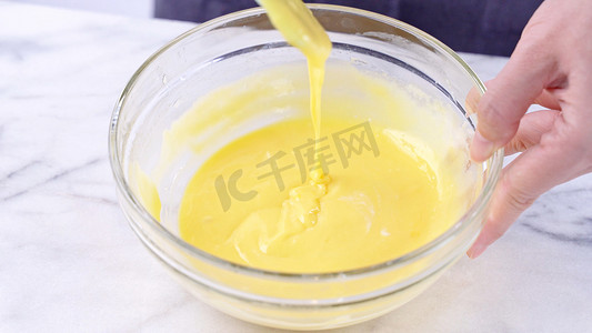 绑带雪纺衫摄影照片_用绿色橡胶刮刀搅拌工具将蛋黄混合到蛋糕面糊中，搅拌至光滑，并在玻璃碗中充分混合，特写，生活方式