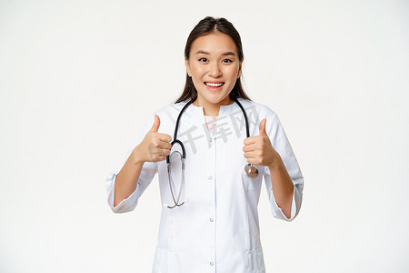 微笑的亚洲女医生，穿着医用长袍，高兴地竖起大拇指，推荐保健品，站在白色背景上