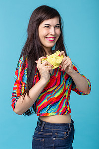 微笑的年轻女子手里拿着一个摆在蓝色背景上的黄色老式相机模型。