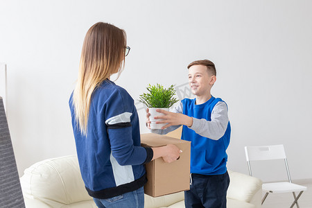 单身摄影照片_年轻的单身母亲和一个顽皮的小儿子站在新公寓的客厅里，手里拿着一个盒子，里面装着东西，花盆里放着一朵花。