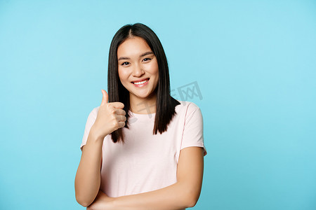 美丽的微笑亚洲女孩竖起大拇指，赞同，同意，推荐公司品牌，站在蓝色背景上