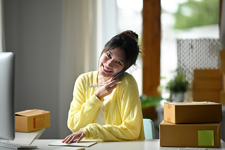 微笑的女性在线卖家使用电脑和手机对话来确认客户的订单。