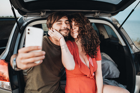 幸福的白人情侣男人和卷发黑发女人坐在 SUV 车的后备箱里用智能手机自拍