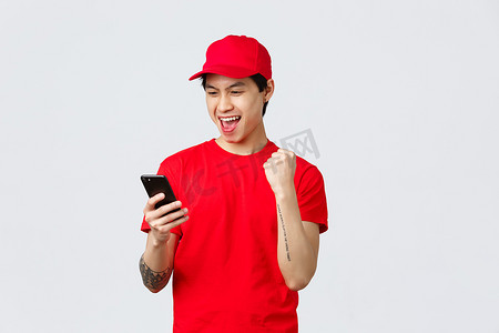 身穿红色制服 T 恤和帽子的快乐的年轻亚洲送货员，阅读好消息智能手机屏幕，挥拳高呼巨大的奖金或成功。