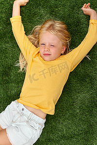 肖像可爱的金发女孩闭着眼睛躺在绿色的草地上伸展。