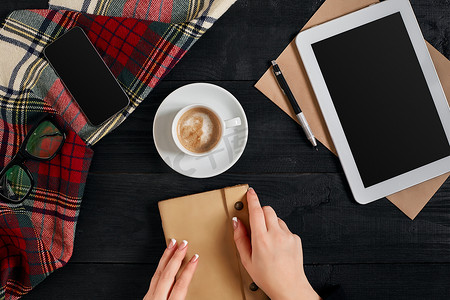早上，年轻女性右手在木桌上的空白笔记本上写字，旁边放着咖啡杯、智能手机和眼镜