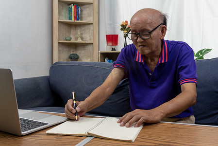 留言条摄影照片_亚洲老人拿着笔在沙发上的桌子上写留言条和电脑笔记本电脑。