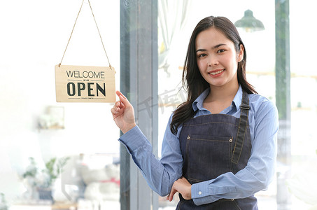 亚洲快乐的女商人是一位穿着围裙的女服务员，咖啡馆的老板站在门口，上面挂着“开放”的牌子等待顾客。