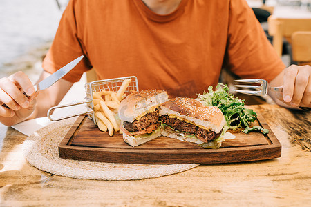 在街边咖啡馆的盘子里，坐在切片烤美味牛肉奶酪汉堡包和炸薯条前的裁剪特写男性男性。