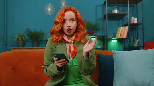 巨大手机摄影照片_在家沙发上的年轻红发女孩使用智能手机打字浏览说哇是发现了巨大的胜利