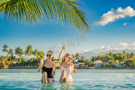 暑假期间，两个美丽的年轻女子坐在度假村游泳池的池畔。