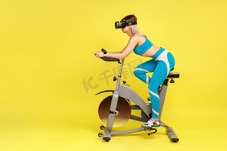 苗条女子戴着虚拟现实耳机骑健身自行车进行有氧运动的肖像。