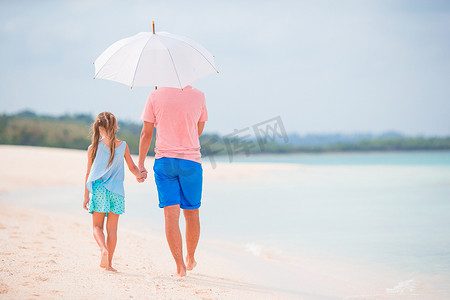 年轻的父亲和他的小女儿在白色沙滩上的伞下行走