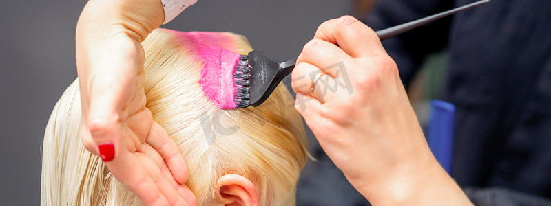 美发沙龙摄影照片_在美发沙龙中，用刷子在一位年轻金发女子的白发上涂上粉红色染料。
