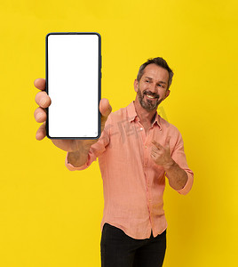 巨大手机摄影照片_快乐的中年灰发健壮的男人拿着大智能手机指着它，穿着与黄色隔离的桃色衬衫。