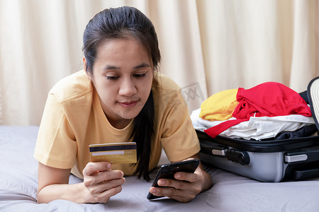 旅游计划摄影照片_亚洲快乐的女人计划在家里的床上用手机和信用卡旅行。