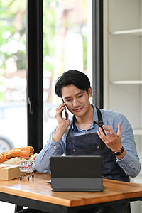 年轻的小企业主使用电脑和手机通话来确认客户的订单。