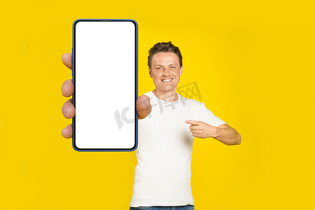 英俊的金发男子指着巨大的智能手机与白色空屏幕，穿着白色 T 恤和牛仔裤手机显示模拟孤立在黄色背景。