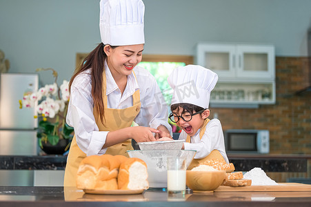 烘焙蛋糕广告图摄影照片_可爱的亚洲小男孩和漂亮的妈妈在桌上的家庭厨房里用筛子筛漏勺筛面粉，准备烘焙面包店和蛋糕。
