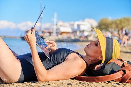 戴着笔记本电脑的帽子在沙滩上晒日光浴的成熟女性