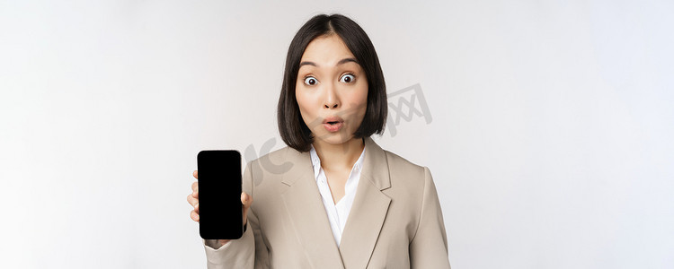 亚洲企业女性的形象显示应用程序界面、手机屏幕、惊讶的表情、哇、站在白色背景上