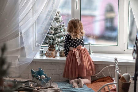 一个小女孩坐在窗边的床上，用圣诞小玩具装饰一棵小树。
