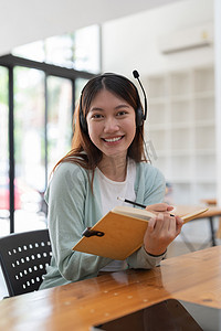 戴着耳机书写的亚洲年轻女性画像在记事本中记笔记，在室内笔记本电脑上工作或学习 — 教育课程或培训、研讨会、在线教育概念