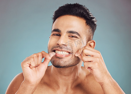 一位微笑的年轻印度男子在蓝色工作室背景下用牙线清洁牙齿的肖像。