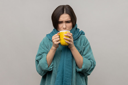 围巾摄影照片_戴围巾的病态不健康妇女手握热茶杯，感觉寒冷，喝热饮料