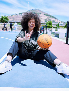 时尚的年轻西班牙裔女性用双手和手指做出摇滚手势，在球场上举着篮球。