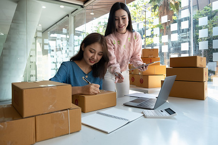 确认订单摄影照片_两名亚洲女员工使用笔记本电脑在仓库工作并浏览在线购物订单文件以确认库存商品。