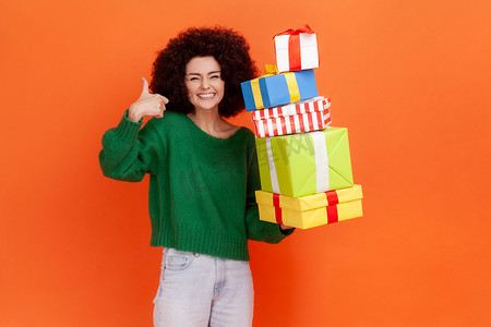 绿色购物节摄影照片_快乐、满意的女人，留着非洲式发型，穿着绿色休闲风格的毛衣，站着很多礼物盒，竖起大拇指，微笑着。