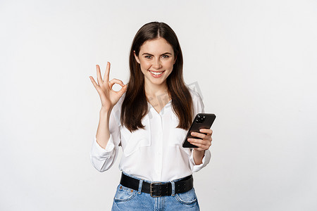 微笑的女人在使用手机时表现得很好，很好的手势，批准智能手机上的应用程序，优质的服务，网上商店，站在白色背景上