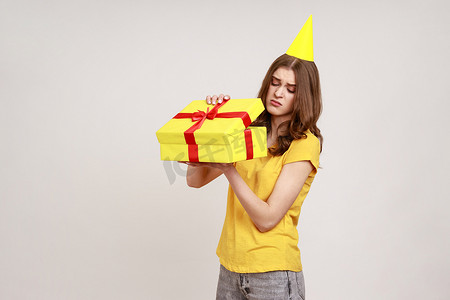 心烦意乱的少女打开礼盒，带着失望的表情看着相机，打开坏礼物，穿着黄色 T 恤和派对锥。