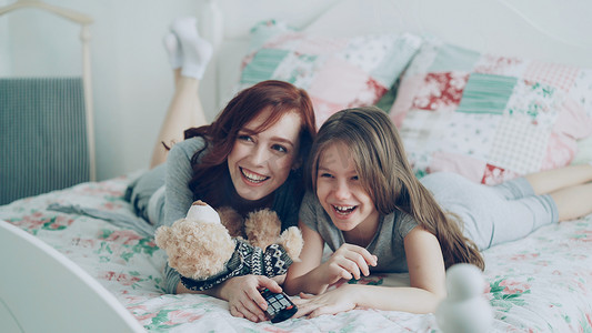 开心的女孩卡通摄影照片_早上，快乐的妈妈和可爱的女儿躺在家里的床上，看着电视上有趣的卡通电影，玩得很开心