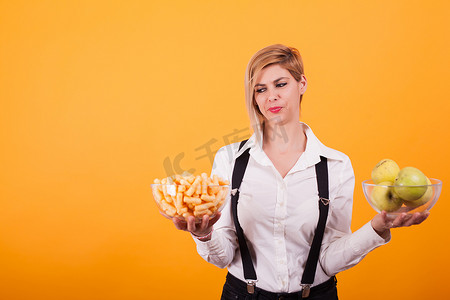 年轻漂亮的女人拿着两碗玉米泡芙和青苹果，背景是黄色的。