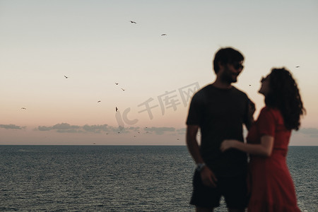 甜水面摄影照片_傍晚日落后，一对模糊的夫妇站在大海的背景上，海鸥飞过水面