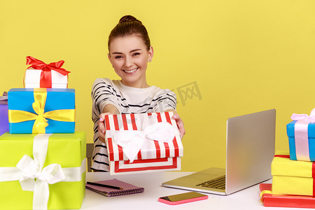 女人坐在工作场所，拿着笔记本电脑，拿着礼品盒，给相机送礼物，祝贺。