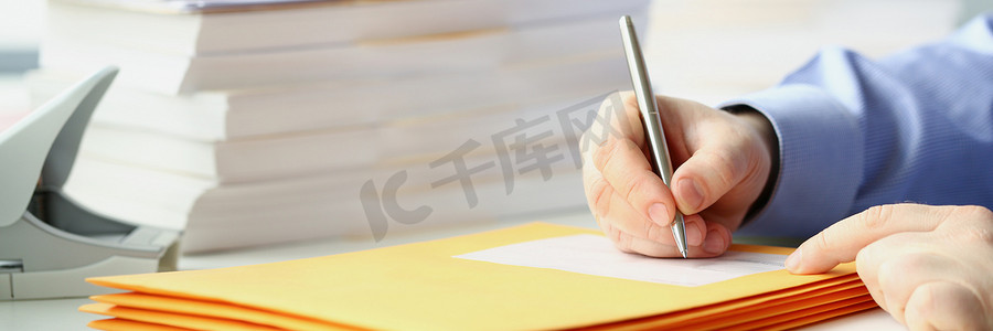拿笔的手摄影照片_手拿笔在信封上签名的男人坐在桌旁，手里拿着一堆文件