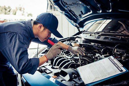 汽车修理师在汽车修理店车库维修服务中按客户索赔订单拿着带检查表剪贴板的手电筒检查发动机来维修车辆。