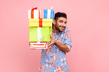 积极的男人的肖像，露出露齿的微笑，拿着一堆礼物，看着相机，送礼物，穿着蓝色休闲风格的衬衫。