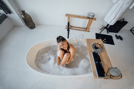 洗澡女人摄影照片_坐在浴缸里的年轻女人抱着双腿