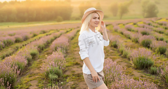 美丽的年轻健康女人，穿着白色连衣裙，在夕阳的光芒下拿着草帽，快乐地穿过薰衣草田奔跑