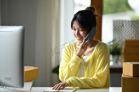 微笑的女性小企业主使用电脑和手机通话来确认客户的订单。