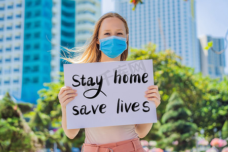 戴医用口罩的妇女可预防冠状病毒疾病，她持有一张海报“呆在家里拯救生命”手写文字 — 字母被隔离在白色上。
